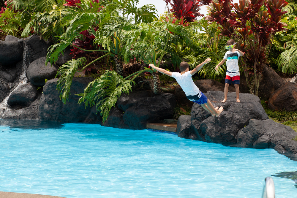Photo of boy falling backward into pool of water, boy wearing goggles stands on rock looking on