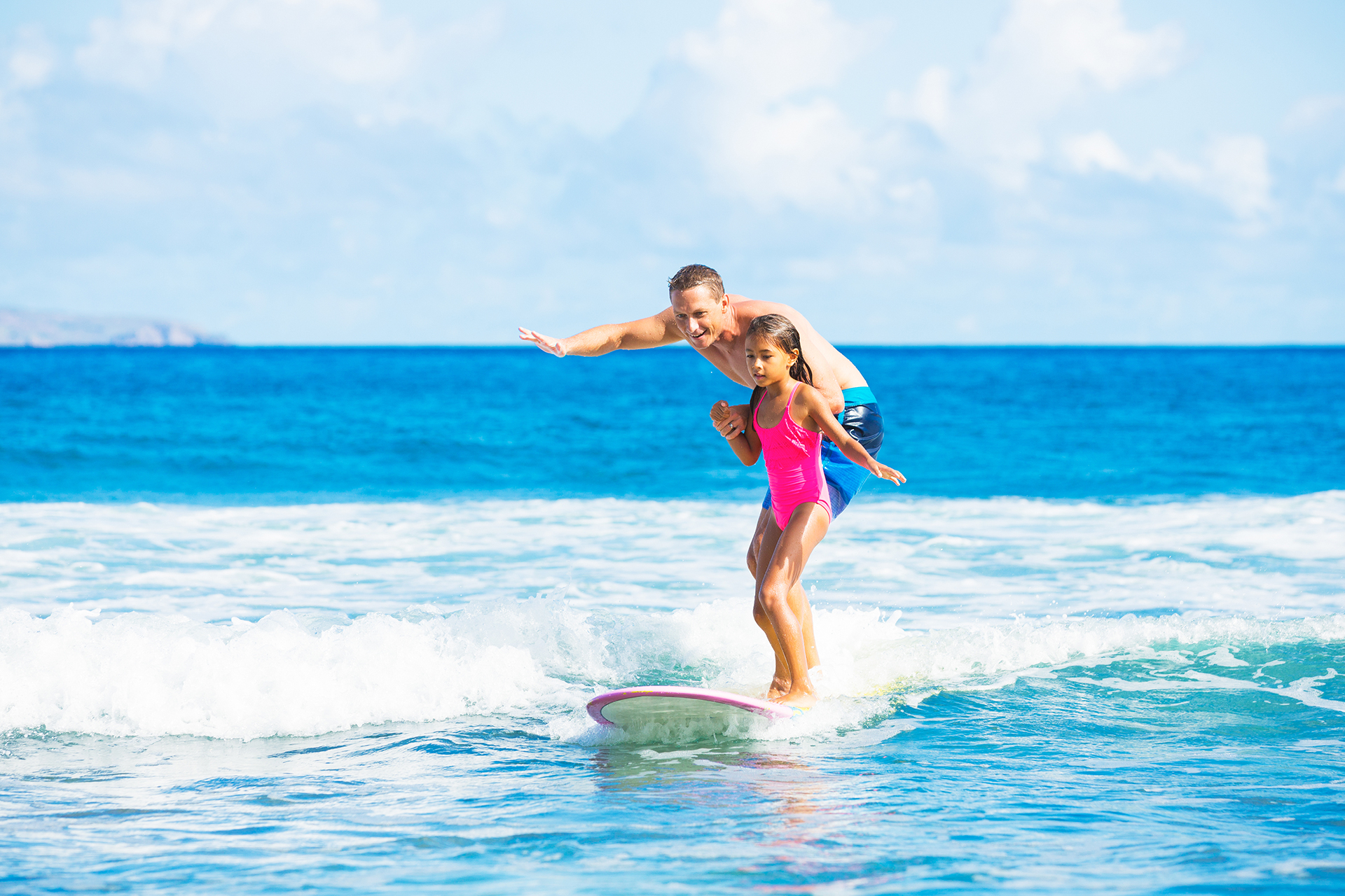 Girl in fluorescent pink bathing suit on surfboard, with adult male behind her, coaching her to surf