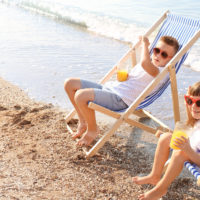 Two children drinking juice on the beach in beach chairs