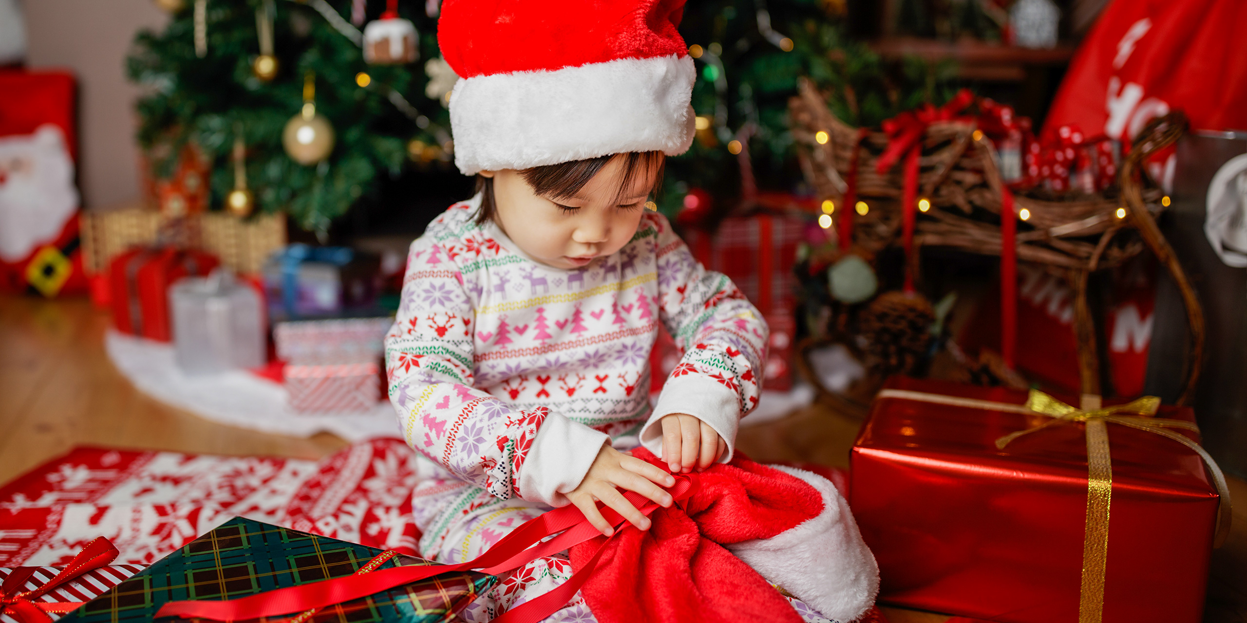toddler baby girl wearing santa claus costume looking for gift in front of christmas tree - Image ; Mcimage /Shutterstock