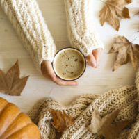 Aerial view of hands in white sweater holding coffee while surrounded by pumpkins and leaves