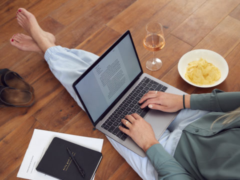 Woman typing on computer next to plate of food and glass of wine, wearing loungewear pants