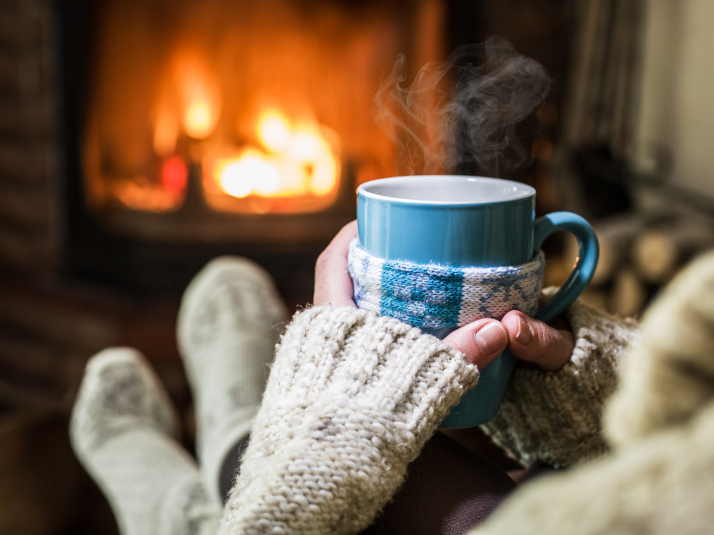 Person holding a steaming mug in front of a lit fireplace
