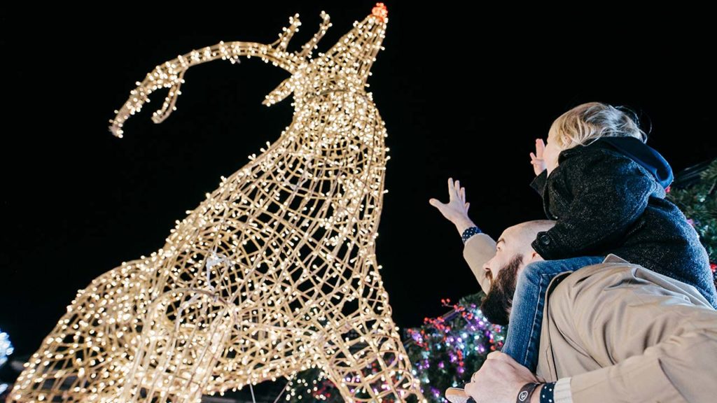 Small child on father's shoulders reaching up toward a light up reindeer sculpture