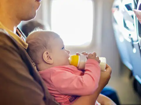 Father holding his baby daughter during flight on airplane going on vacations. Baby girl drinking formula milk from bottle. ; Courtesy of Romrodphoto/Shutterstock