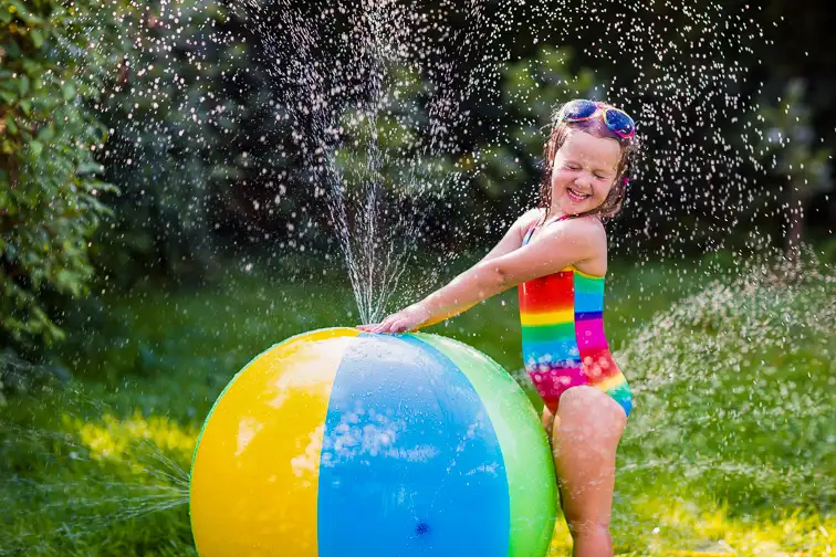 Funny laughing little girl in a colorful swimming suit playing with toy ball garden sprinkler with water splashes having fun in the backyard on a sunny hot summer vacation day. ; Courtesy of Famveld/Shutterstock