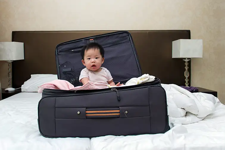 Asian baby sitting in travel suitcase placed on hotel double bed; Courtesy of Kenishirotie/Shutterstock
