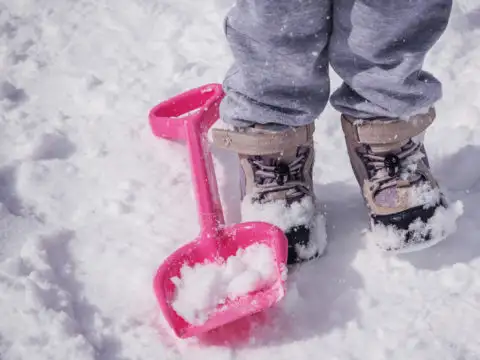 Child wearing snow boots in the snow next to a snow toy shovel