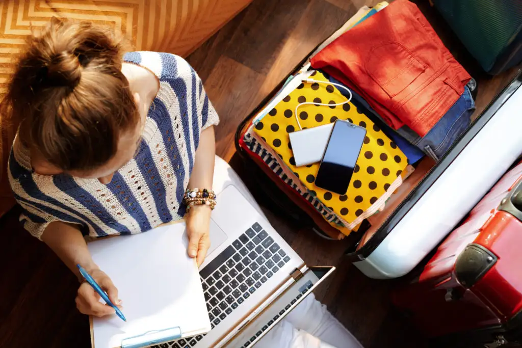 Woman sitting next to a packed suitcase, planning a vacation with a laptop and a notebook