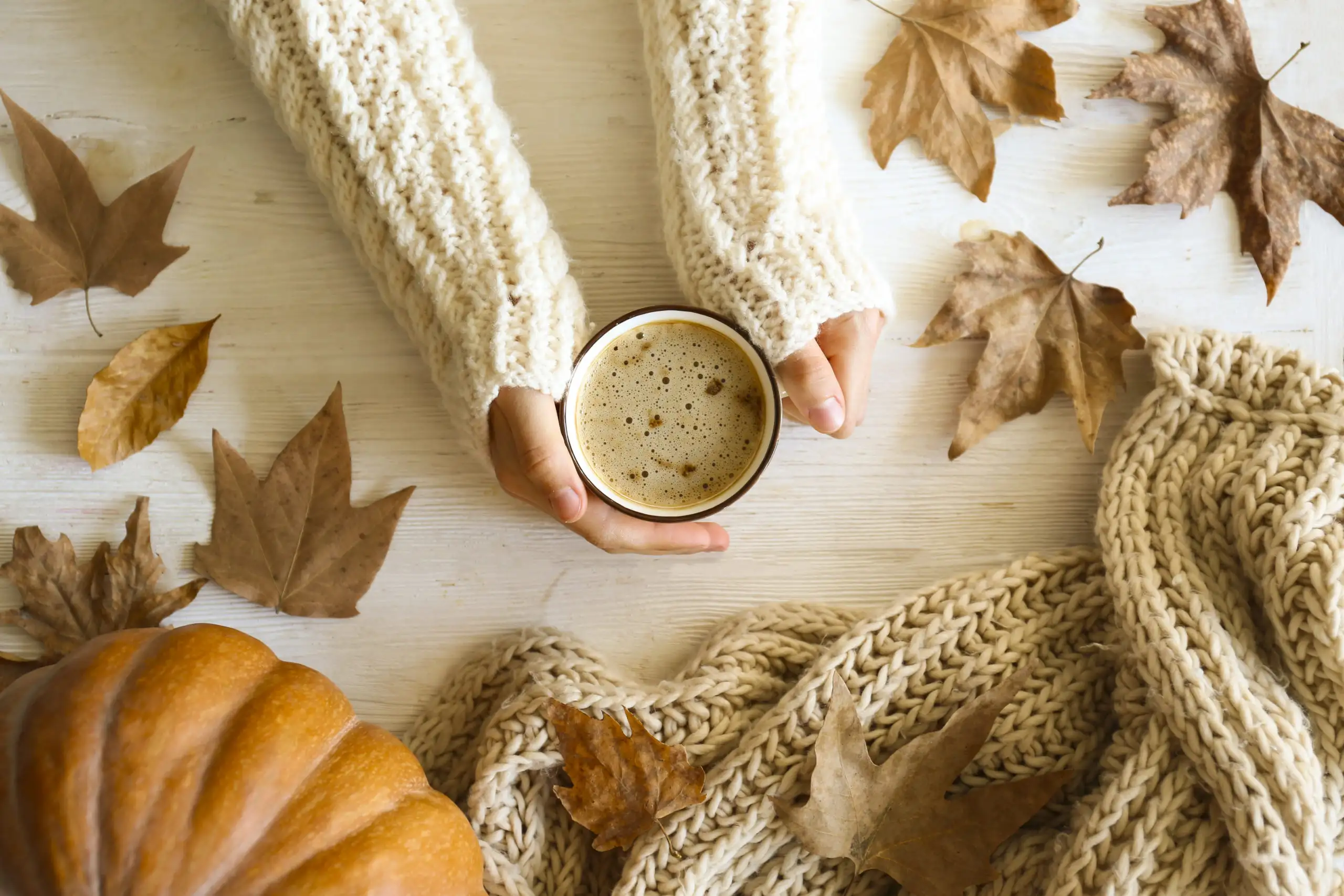 Aerial view of hands in white sweater holding coffee while surrounded by pumpkins and leaves