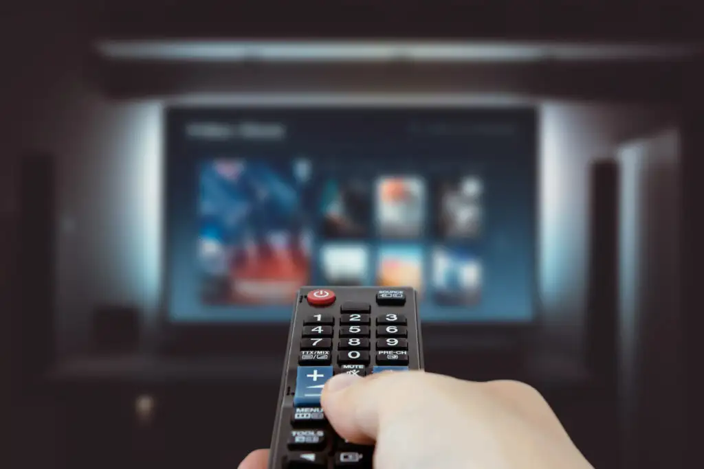 Hand holding TV remote in the foreground with blurry TV in the background