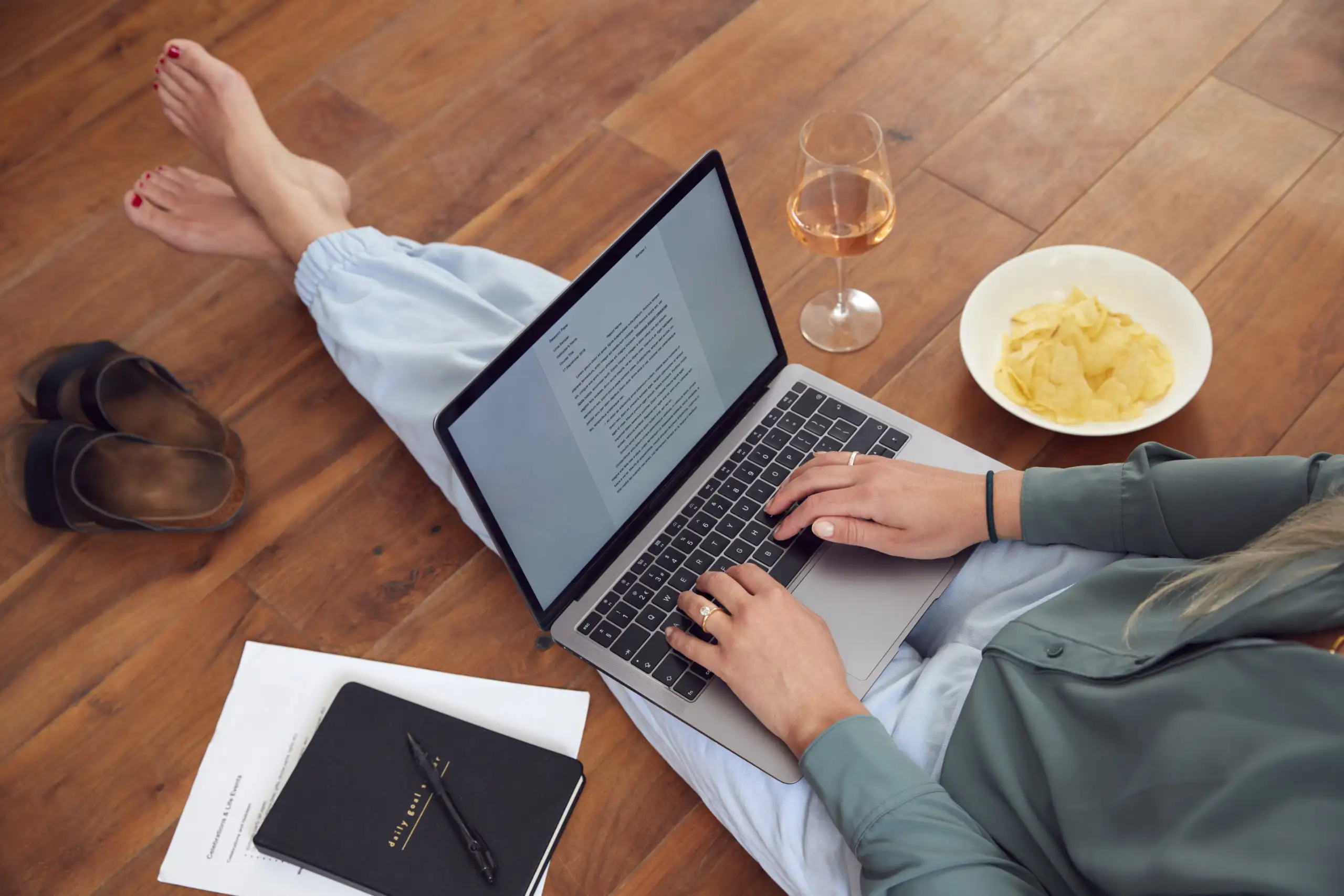 Woman typing on computer next to plate of food and glass of wine, wearing loungewear pants