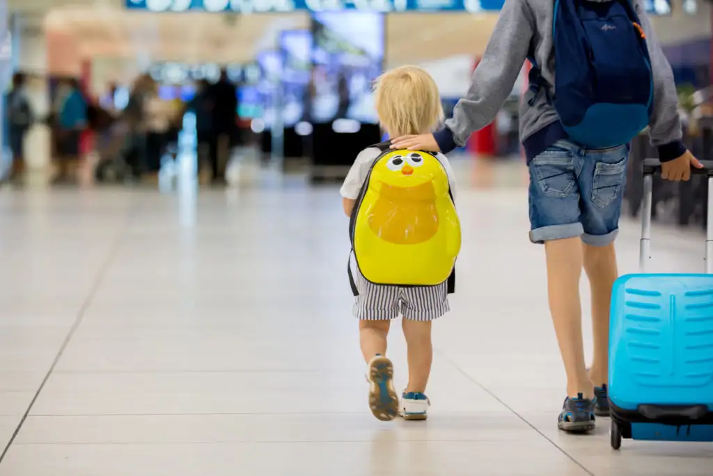 Young child walking through airport with a yellow backpack, guided by older child with a blue rolling suitcase