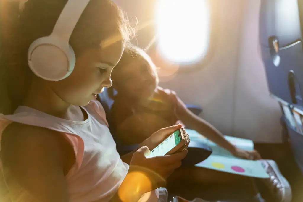 Child wearing over-ear headphones while playing on phone on plane with second child looking at screen from next seat over